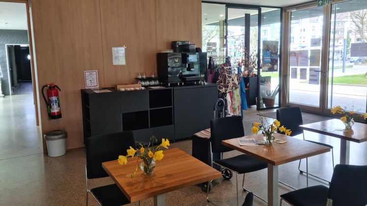 Innenraum des Gemeindezentrums mit Tischen, Stühlen, Kaffeemaschine und Souvenirladen.