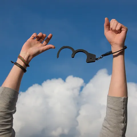 Hände gegen blauen Himmel erhoben, ein Handgelenk befreit von zerbrochenen Handschellen.