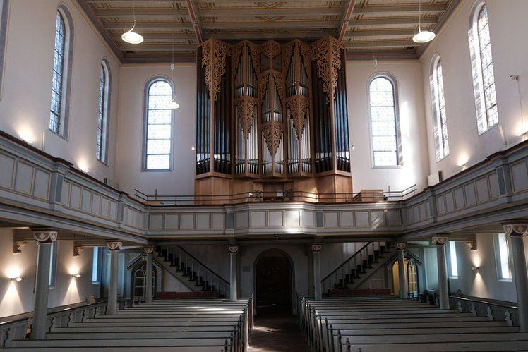 Kircheninneres mit Pfeifenorgel, Bänken, Emporen und Bogenfenstern.