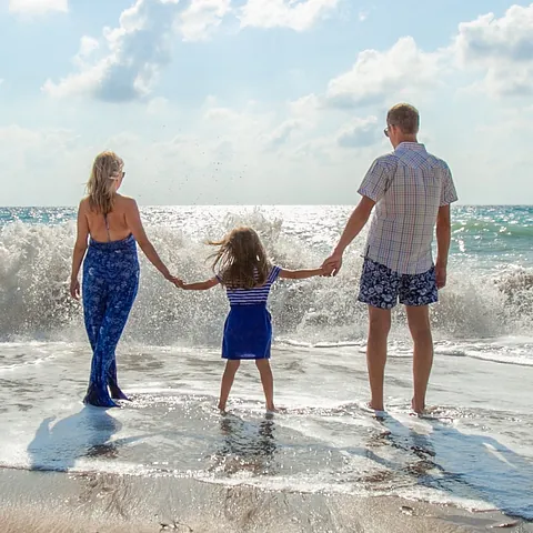 Dreiköpfige Familie hält Händchen und blickt auf Meereswellen an einem sonnigen Strand.
