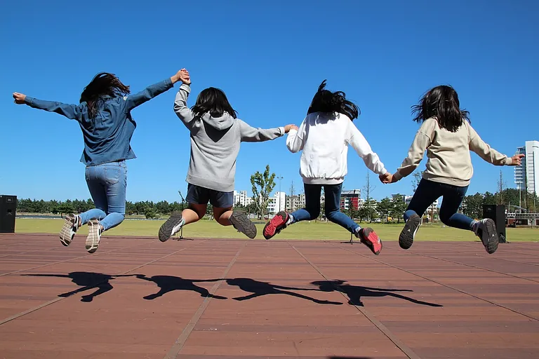 Vier junge Frauen halten Händchen und springen fröhlich unter blauem Himmel.
