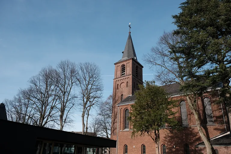 Backsteinkirchturm ragt in klaren blauen Himmel zwischen kahlen Bäumen.