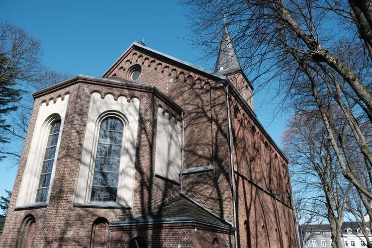 Backsteinkirchenfassade mit Bogenfenstern und Kirchturmspitze vor blauem Himmel.