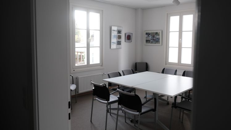 Kleiner Besprechungsraum mit weißem Tisch, Stühlen und Tageslicht.