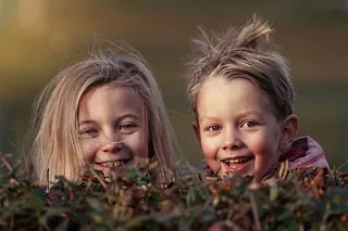 Zwei fröhliche junge Kinder schauen über eine grüne Hecke und lächeln hell.