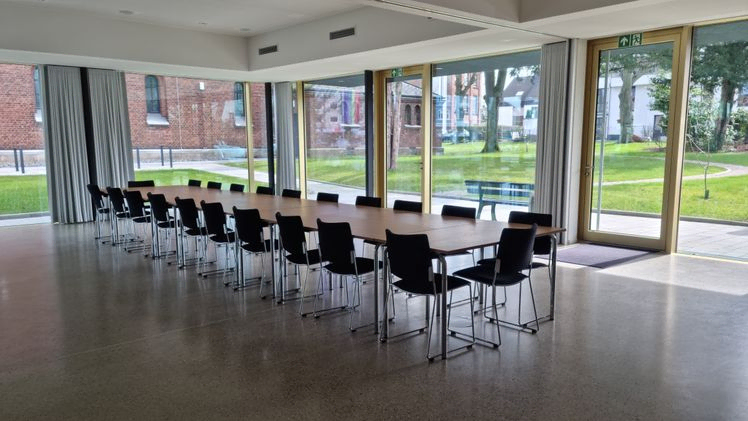 Geräumiger Gemeinschaftssaal mit langen Tischen und Stühlen, Tageslicht.