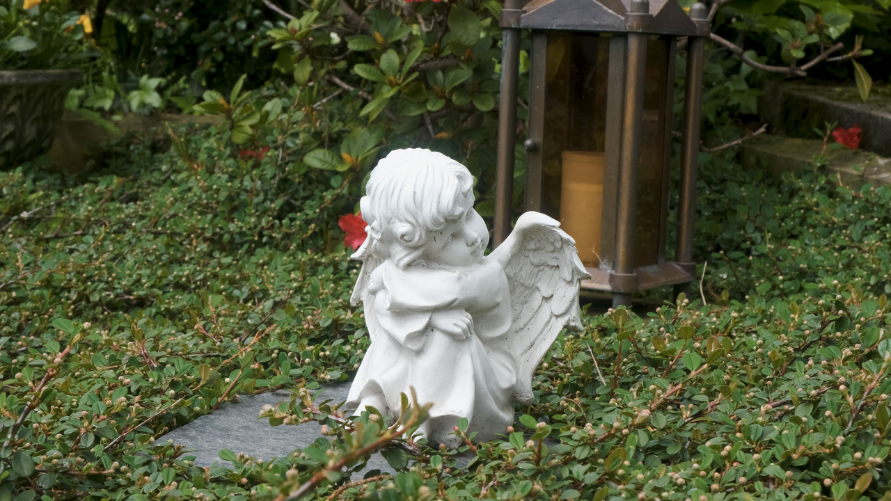 Weiße Engelsstatue sitzt im Grün neben einer Laterne auf einem Grab.